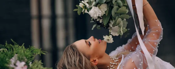 Une mariée allongée, tenant un bouquet de fleurs blanches et vertes, vêtue d'une robe ornée de perles, avec un collier assorti, dans un cadre élégant et contrasté.