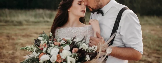 Mariés s'enlaçant tendrement dans un champ au coucher du soleil, la mariée tenant un grand bouquet de fleurs bohèmes.