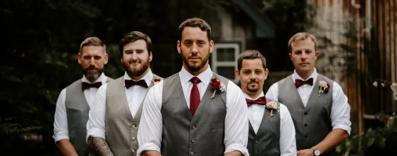 Un groupe de cinq hommes en tenue de mariage, portant des gilets gris, des chemises blanches et des nœuds papillon bordeaux. L'homme au centre porte une cravate assortie et une boutonnière florale. Ils posent de manière formelle en extérieur, avec un fond naturel et rustique.