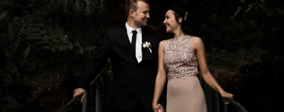 Un couple élégamment habillé se tient la main sur un petit pont en bois, entouré d'une végétation dense. L'homme porte un costume noir avec une cravate et une boutonnière blanche, tandis que la femme est vêtue d'une robe longue rose pâle avec un haut scintillant. Ils se regardent avec complicité.
