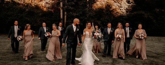 Un couple de mariés marche main dans la main avec leur cortège nuptial dans un cadre naturel au coucher du soleil.