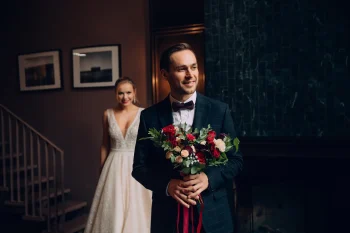 Un marié souriant tenant un bouquet de fleurs, tandis que la mariée s’approche de lui en arrière-plan avec un sourire radieux.