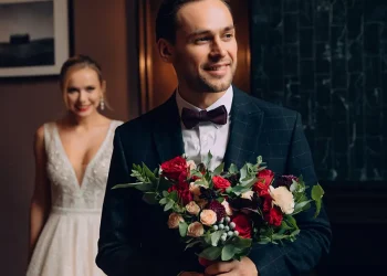 Un homme élégant en costume sombre avec un nœud papillon tient un bouquet de fleurs aux couleurs vives, tandis qu’une femme en robe de mariée blanche s’approche en arrière-plan avec un sourire radieux.