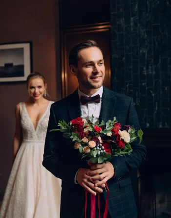 Un homme élégant en costume sombre avec un nœud papillon tient un bouquet de fleurs aux couleurs vives, tandis qu’une femme en robe de mariée blanche s’approche en arrière-plan avec un sourire radieux.