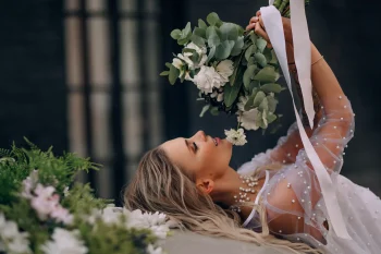 Une mariée allongée, tenant un bouquet de fleurs blanches et vertes, vêtue d'une robe ornée de perles, avec un collier assorti, dans un cadre élégant et contrasté.