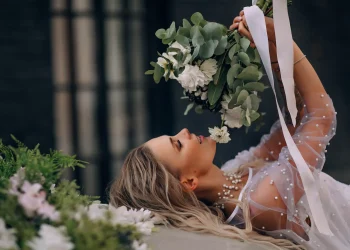 Une mariée allongée, tenant un bouquet de fleurs blanches et vertes, vêtue d'une robe ornée de perles, avec un collier assorti, dans un cadre élégant et contrasté.