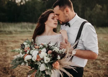 Mariés s'enlaçant tendrement dans un champ au coucher du soleil, la mariée tenant un grand bouquet de fleurs bohèmes.