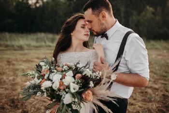 Mariés s'enlaçant tendrement dans un champ au coucher du soleil, la mariée tenant un grand bouquet de fleurs bohèmes.