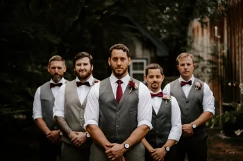 Un groupe de cinq hommes en tenue de mariage, portant des gilets gris, des chemises blanches et des nœuds papillon bordeaux. L'homme au centre porte une cravate assortie et une boutonnière florale. Ils posent de manière formelle en extérieur, avec un fond naturel et rustique.