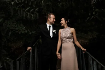 Un couple élégamment habillé se tient la main sur un petit pont en bois, entouré d'une végétation dense. L'homme porte un costume noir avec une cravate et une boutonnière blanche, tandis que la femme est vêtue d'une robe longue rose pâle avec un haut scintillant. Ils se regardent avec complicité.