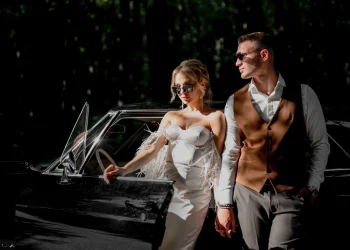 Un couple élégant pose près d'une voiture vintage noire en pleine nature. La femme porte une robe blanche bustier avec des détails en plumes et des lunettes de soleil. L'homme est vêtu d'une chemise blanche, d'un gilet marron et de lunettes de soleil. Ils se tiennent par la main, dégageant une ambiance chic et cinématographique.