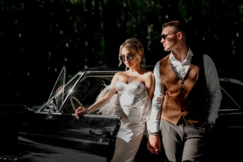 Un couple élégant pose près d'une voiture vintage noire en pleine nature. La femme porte une robe blanche bustier avec des détails en plumes et des lunettes de soleil. L'homme est vêtu d'une chemise blanche, d'un gilet marron et de lunettes de soleil. Ils se tiennent par la main, dégageant une ambiance chic et cinématographique.