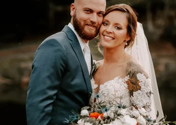 Un couple de mariés sourit avec complicité, la mariée tenant un bouquet de fleurs blanches et orangées, dans un cadre naturel et chaleureux.