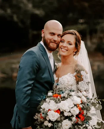 Un couple de mariés sourit avec complicité, la mariée tenant un bouquet de fleurs blanches et orangées, dans un cadre naturel et chaleureux.