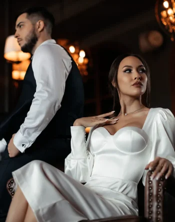 Mariée en robe blanche élégante assise sur un fauteuil, regardant sur le côté, tandis que le marié, en costume noir et chemise blanche, est de dos dans un cadre chaleureux et raffiné
