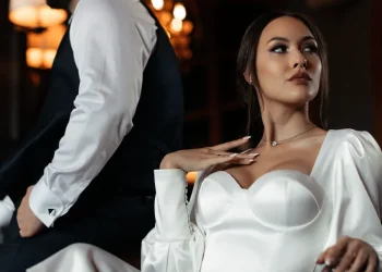 Mariée en robe blanche élégante assise sur un fauteuil, regardant sur le côté, tandis que le marié, en costume noir et chemise blanche, est de dos dans un cadre chaleureux et raffiné