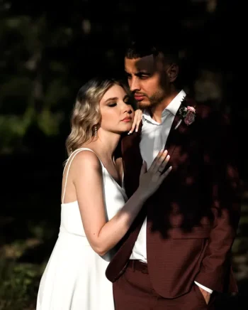 Un couple en tenue de mariage pose tendrement sous une lumière dorée. La mariée, en robe blanche fluide, repose sa tête contre l’épaule du marié, qui porte un costume bordeaux avec une boutonnière florale. Ils sont entourés d'une nature sombre et contrastée.