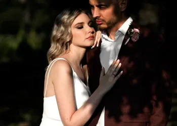 Un couple en tenue de mariage pose tendrement sous une lumière dorée. La mariée, en robe blanche fluide, repose sa tête contre l’épaule du marié, qui porte un costume bordeaux avec une boutonnière florale. Ils sont entourés d'une nature sombre et contrastée.