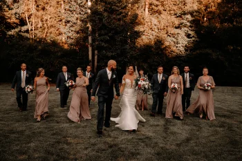 Un couple de mariés marche main dans la main avec leur cortège nuptial dans un cadre naturel au coucher du soleil.